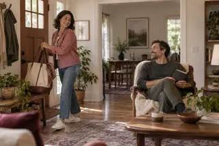 A woman picks up her tote by the front door while a man smiles from a reading chair.