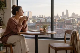 A woman with a mug beside a window and an empty chair.