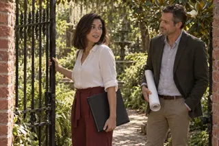 A woman carrying a portfolio opens an iron garden gate while a man with rolled plans waits nearby.