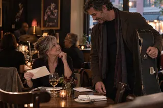 A seated woman holding an envelope looks up at a man carrying an instrument case in a restaurant.