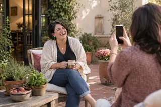A woman laughs in a courtyard while a friend photographs her with a phone.