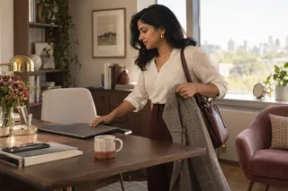 A woman closes her laptop and picks up a jacket beside a sunlit desk.