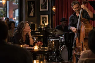 A woman watches a man play double bass beside her table in a warmly lit music venue.