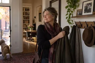 A woman holds a coat beside hallway hooks, with framed photographs above and a dog near the open door.