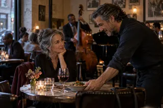 A man sets a dish on a restaurant table as a seated woman looks up at him.