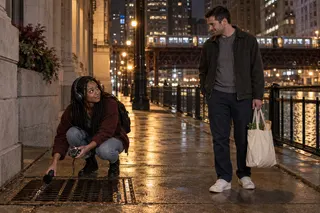 A woman records sound near a storm drain while a man carrying groceries pauses beside her on a nighttime sidewalk.