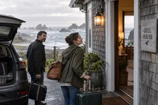 Two adults arrive with luggage at the open doorway of a shingled house overlooking the ocean.