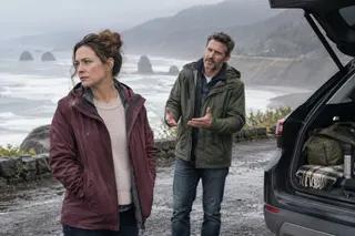 A woman looks away while a man gestures beside an open car trunk at a coastal overlook.