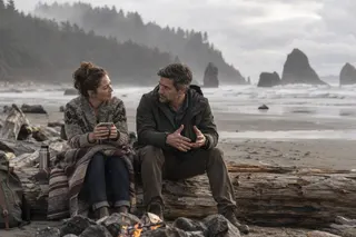 Two adults talk on a driftwood log beside a small beach fire, with a blanket and thermos nearby.