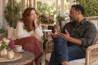 Two adults have an attentive conversation in wicker chairs on a leafy porch.