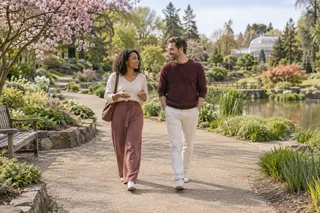 Two adults walk and talk along a garden path beside a pond.