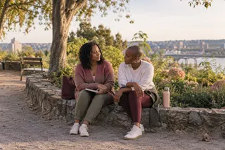 Two women sit together on a low stone wall beside a park path.