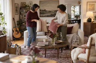 Two women compare cushions beside a sofa in a room with art supplies and a guitar.