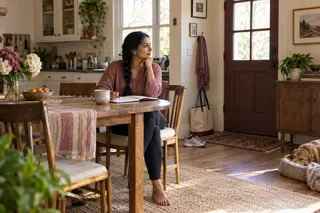 A woman sits with a notebook at a kitchen table, with walking shoes and a tote nearby.
