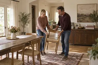 Two adults rearrange chairs around a small dining table in a bright home.