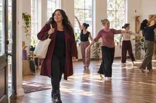 A woman carrying a tote walks through a sunlit dance studio while adults practice behind her.