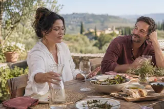 A woman reaches for a water carafe at an outdoor lunch table while another adult listens.