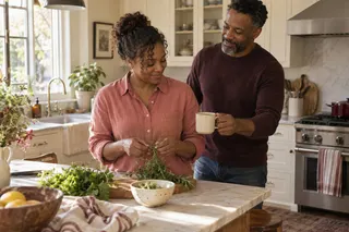 A man offers a mug to a woman sorting fresh herbs at a kitchen counter.
