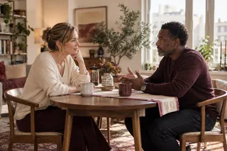 Two adults have a thoughtful conversation across a dining table with mugs of tea.