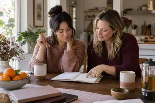 Two women sit together with an open notebook at a breakfast table.
